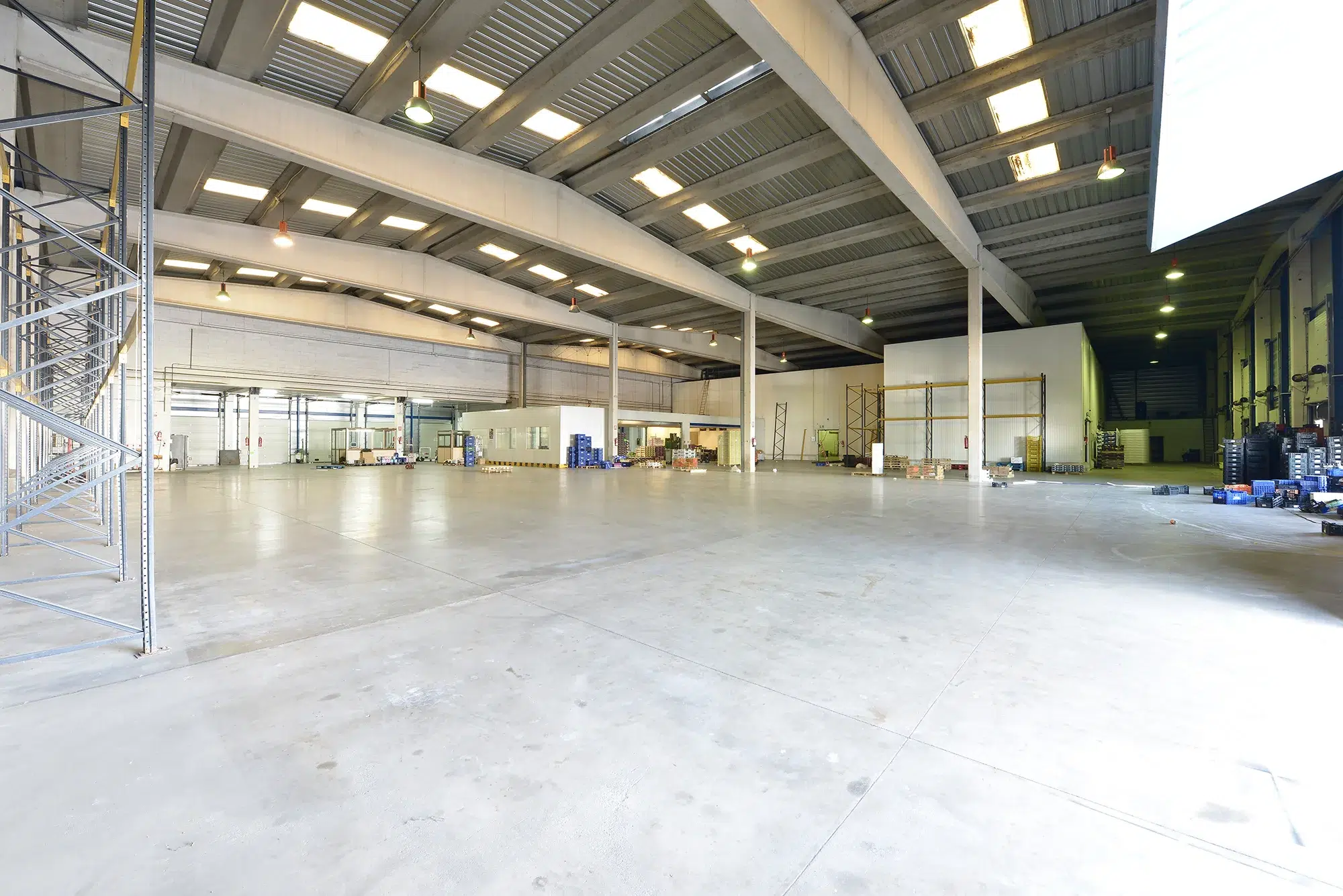 A large empty industrial warehouse with concrete floors and skylights in the ceilings and concrete beams supporting them and metal skeleton shelves